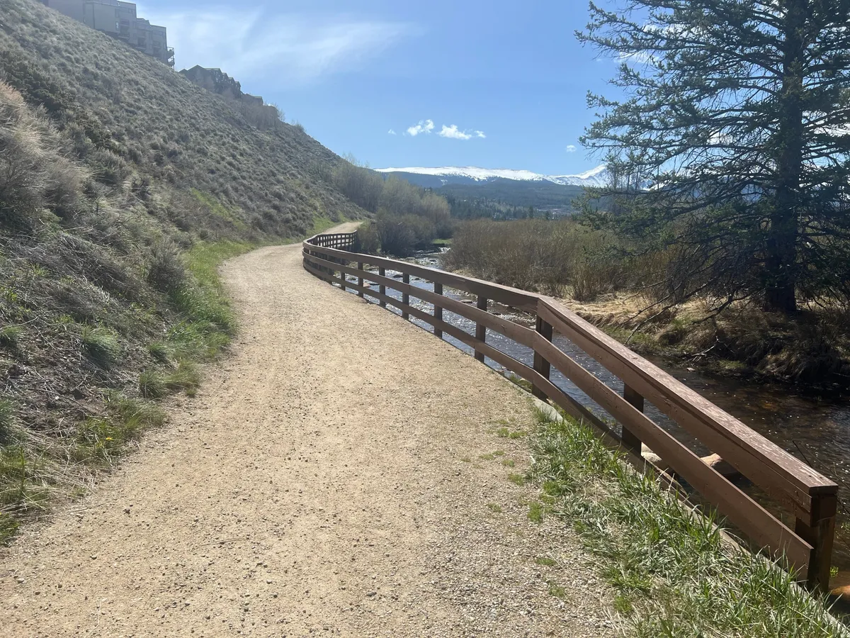 A serene outdoor scene with a winding path, wooden railings, flowing stream, and distant mountain vistas surrounded by lush vegetation. The view evokes peace and connection with nature, inviting exploration and relaxation.