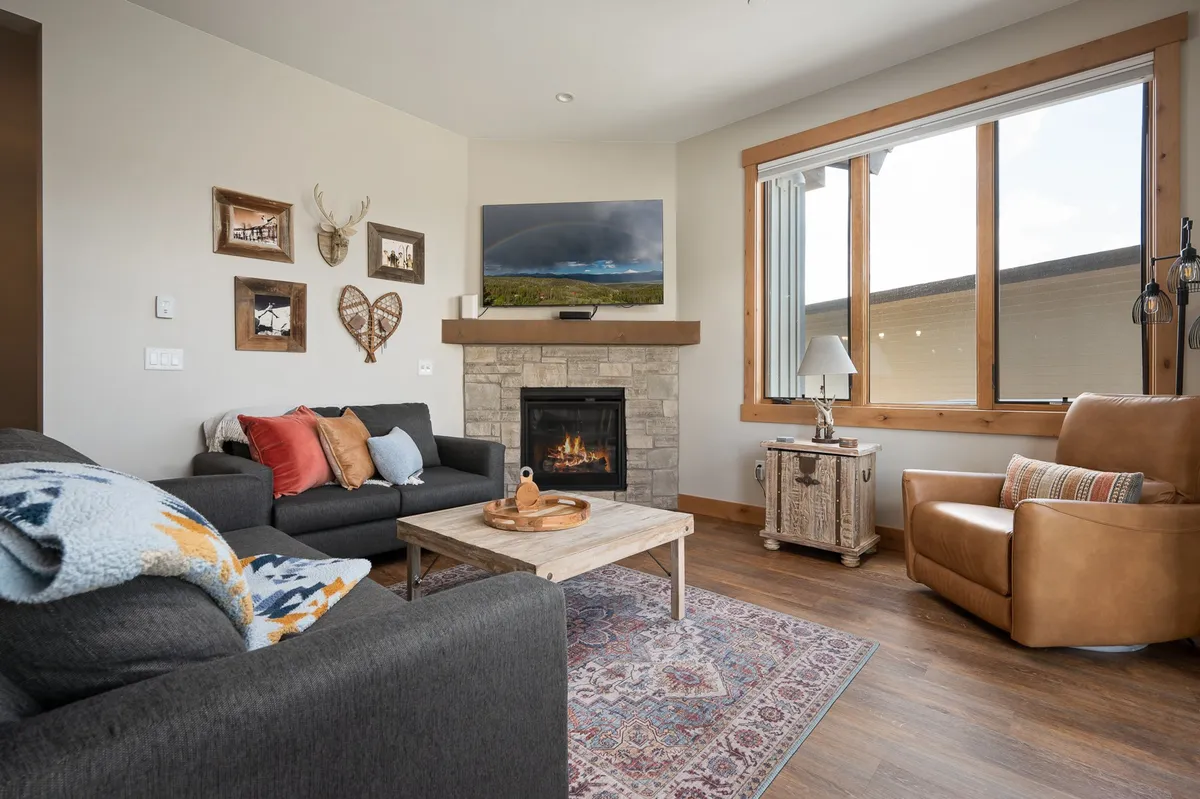 A warm, inviting living room featuring a plush sofa and armchairs arranged around a modern gas fireplace. A coffee table sits at the center, while large windows fill the room with natural light and offer glimpses of the mountain scenery beyond. Artwork adds a touch of personality to the space, and the overall mood is one of comfort and togetherness.