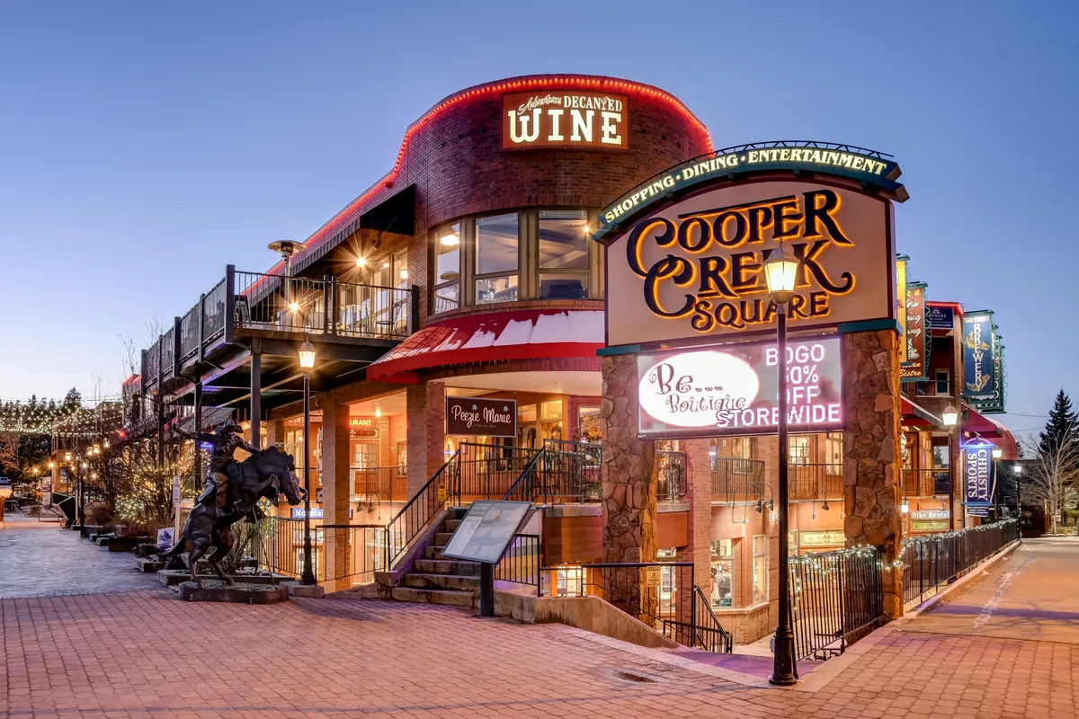 This lively exterior shot shows Cooper Creek Square with its prominent sign, wine shop, upper-level staircase, and a sculpture. The environment is festive and hints at shopping, dining, and community events within walking distance.