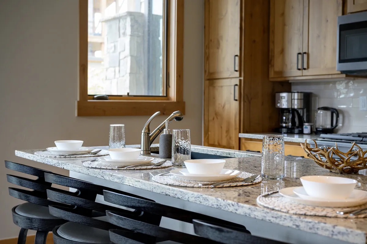 A welcoming kitchen scene with a granite countertop, neatly arranged dishes and glassware, attractive cabinetry, and a window providing natural light. The space is staged for both meal prep and socializing.