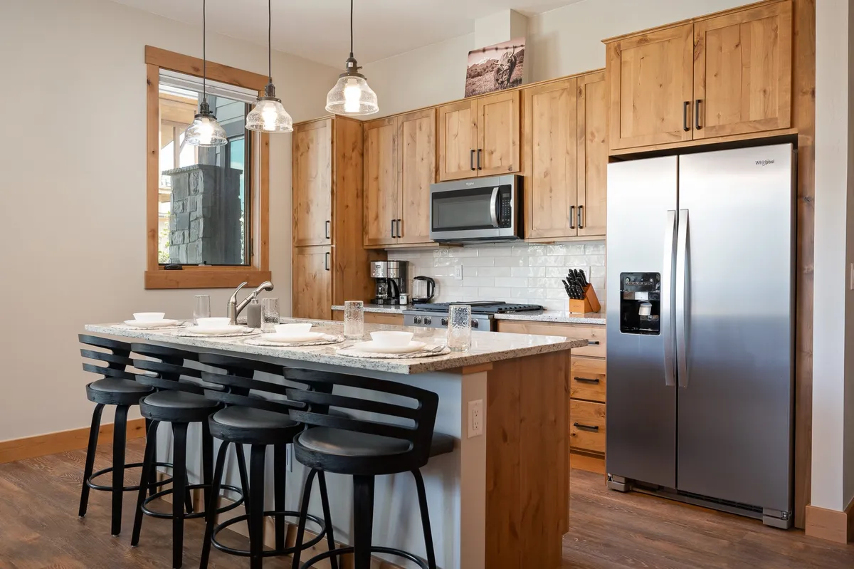 The image portrays a stylish kitchen interior with a large island countertop, gleaming stainless steel appliances, rich wooden cabinetry, and elegant hanging light fixtures. The space is designed for both cooking and entertaining, inviting guests to gather for meals and celebrations in a warm, welcoming atmosphere.
