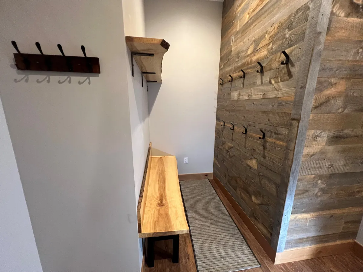 A cozy entryway featuring a wooden bench, a textured wooden wall, and hooks for outdoor gear. The scene suggests a welcoming transition space ideal for returning from outdoor adventures, with practical touches for storing coats and boots.