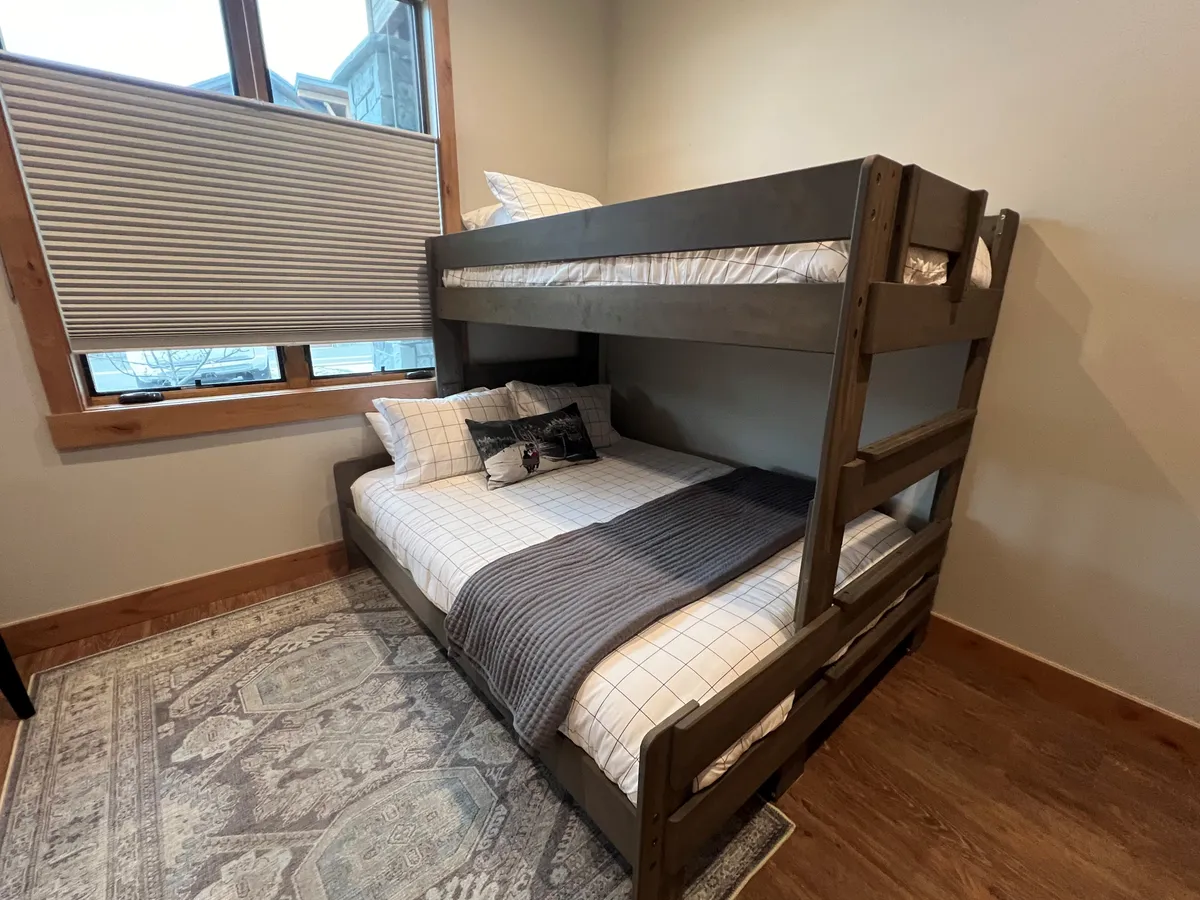 A cozy second bedroom with a twin-over-queen bunk bed, a soft rug, and a sunlit window. The straightforward yet inviting design suggests a flexible and family-friendly sleeping area, ideal for kids or extra guests.