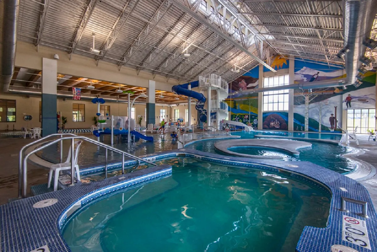 The lively interior of the Grand Park Recreation Center, with children splashing down colorful water slides, families playing in the pool, and parents relaxing poolside. The space is filled with energy, bright murals, and welcoming seating areas.