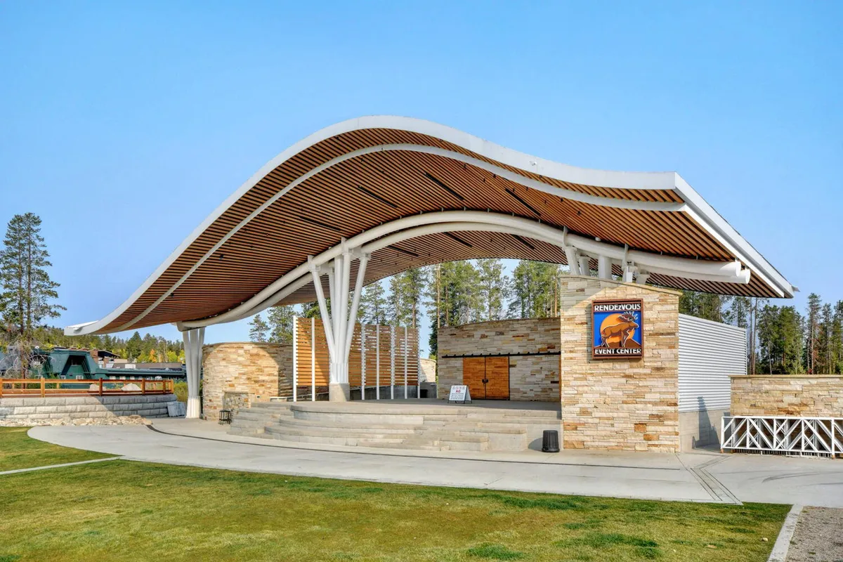 An exterior shot of a curved roof structure with stone walls and wooden accents, set up for community events and concerts. The space is lively, suggesting gatherings, celebrations, and live performances.
