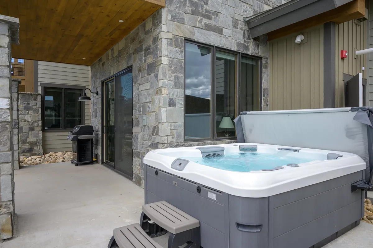 This image highlights an inviting outdoor hot tub set against stone walls, with a grill situated nearby and large windows overlooking the space. The scene captures the tranquil atmosphere of soaking under the open sky, with the patio area ideal for relaxing or gathering with friends, whether during sunny afternoons or star-filled evenings.