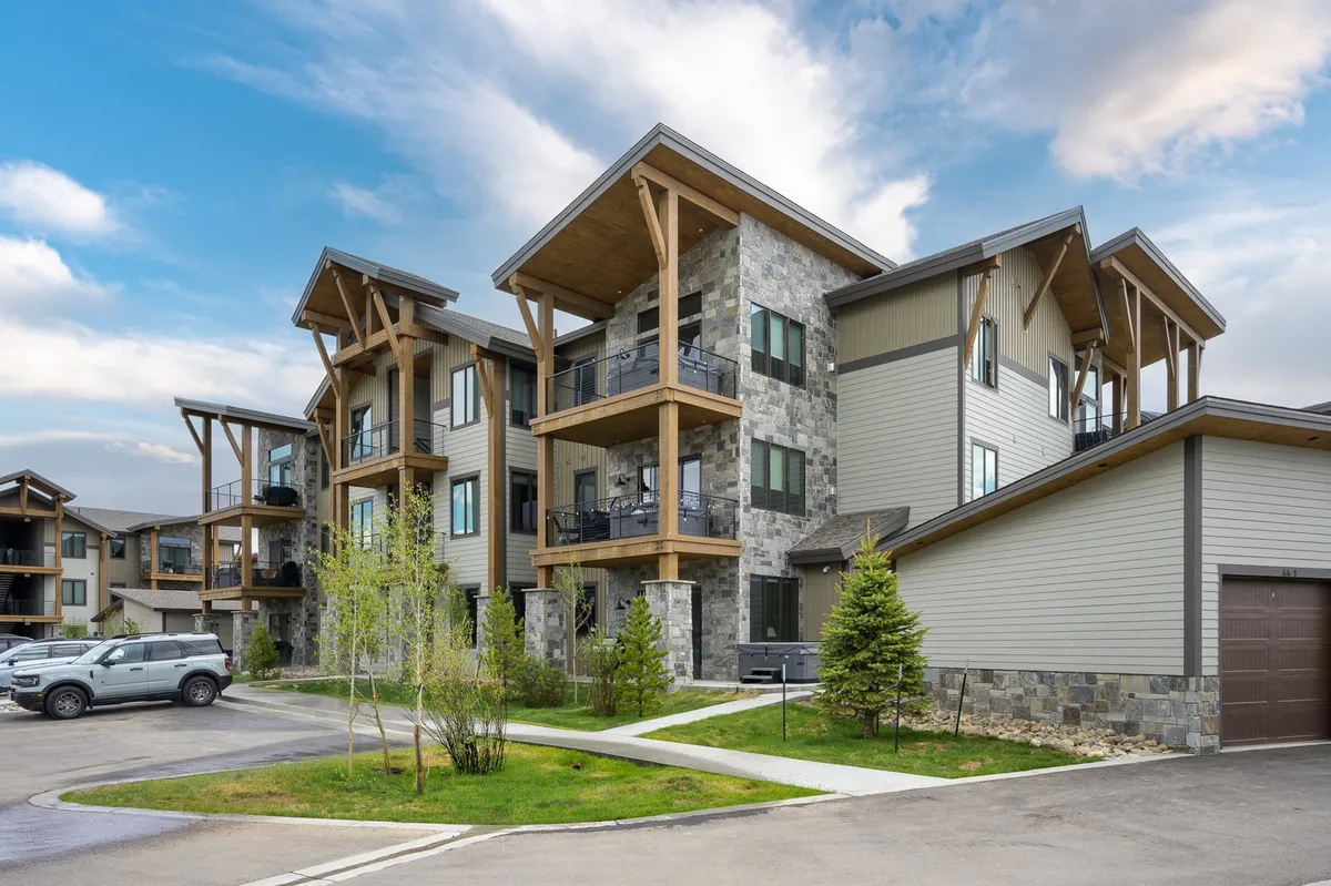 A modern, multi-story building with a stone facade and several balconies, surrounded by lush green landscaping. People can be seen gathered on balconies and in the outdoor areas, suggesting a community atmosphere and the potential for socializing or relaxing outdoors. The image is set during sunset, enhancing the welcoming ambiance.