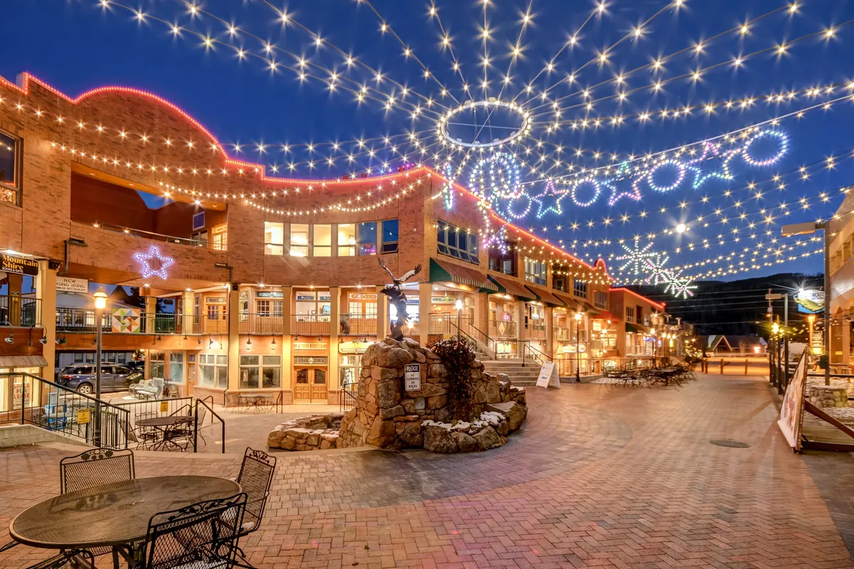 A lively outdoor plaza featuring decorative string lights and a stone fountain. The surrounding architecture suggests a festive, communal setting, ideal for dining, shopping, or attending social events. The evening scene is filled with a celebratory mood, ideal for creating memories with friends or loved ones.